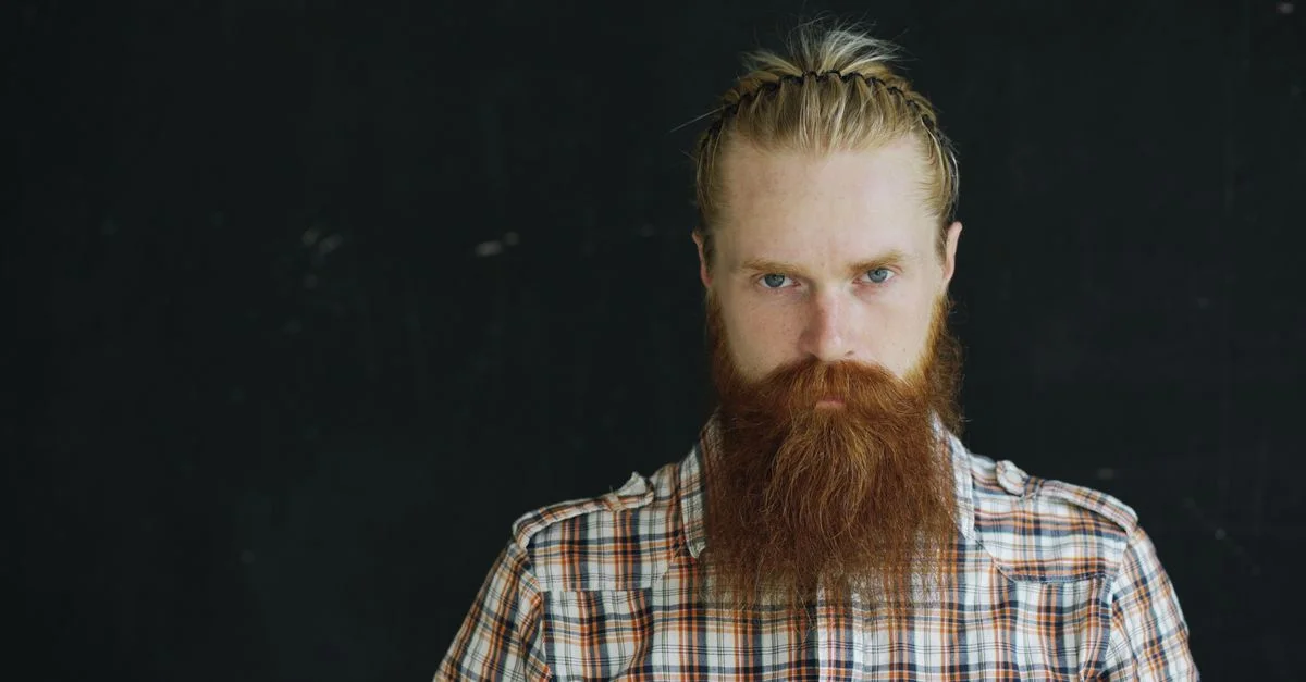 Homme sérieux avec une longue barbe rouge et des cheveux tressés, portant une chemise à carreaux sur fond sombre.